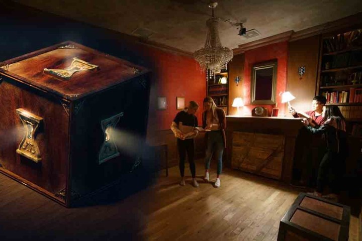 Room with three people solving puzzles and a large wooden cube nearby, featuring a chandelier and bookshelves.