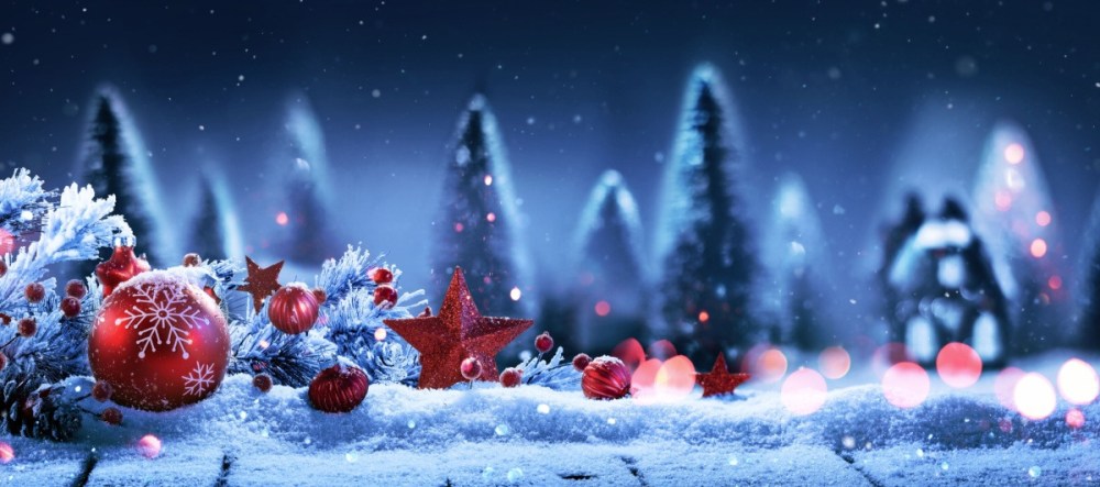 Festive red ornaments and snow on a wooden surface with blurred snowy trees in the background.