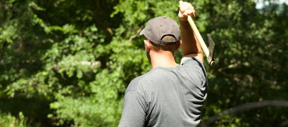 Man in gray shirt and cap holding an axe in the forest.