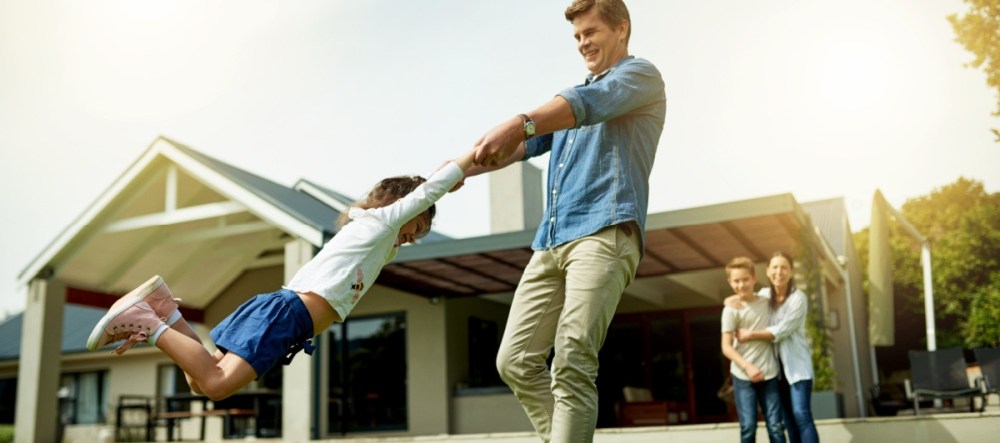 Man spins child in front of modern house with family watching.