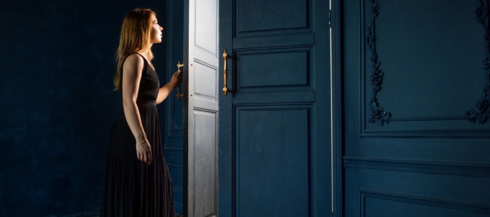 Woman in a black dress opening a blue door to a bright light.