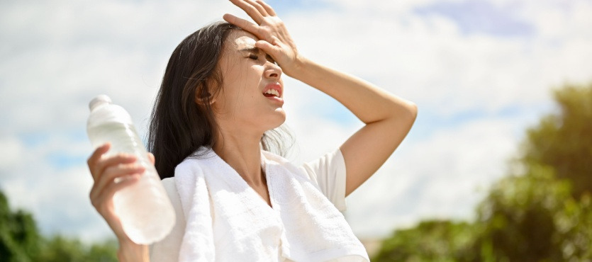 Woman outdoors shielding eyes from sun, holding water bottle, towel around neck.