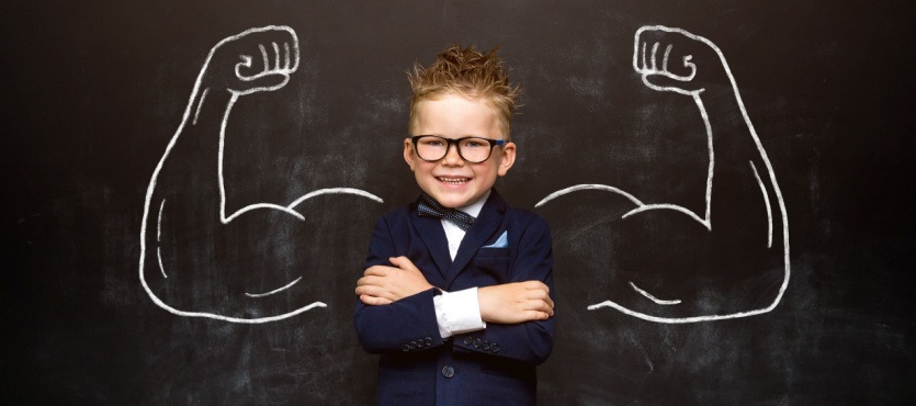 Smiling child in a suit and glasses, arms crossed, with chalk-drawn muscles on a blackboard behind.