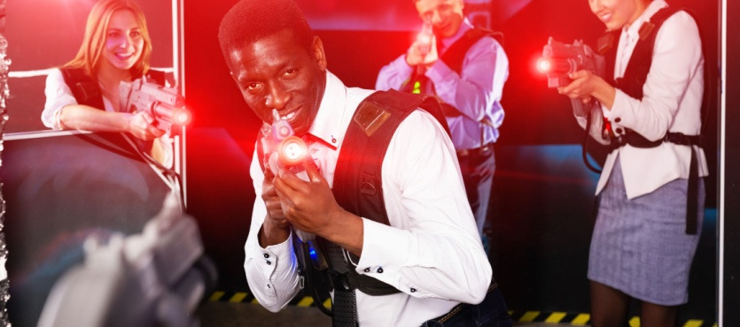 People playing laser tag, aiming at camera with laser guns in a dark arena.