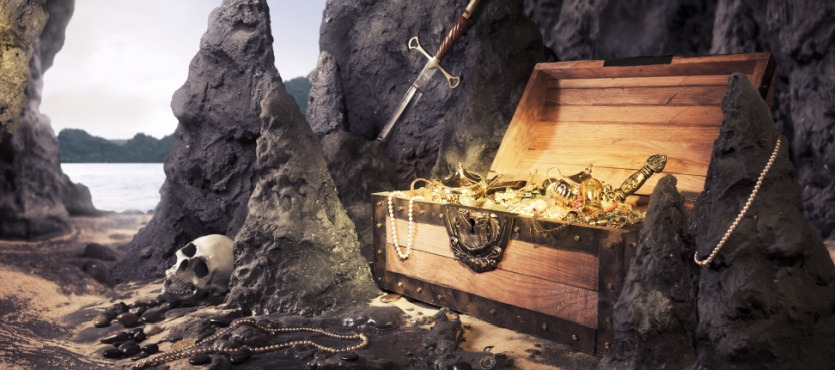 Open treasure chest with jewels, skull, sword, and rocky surroundings on a beach.
