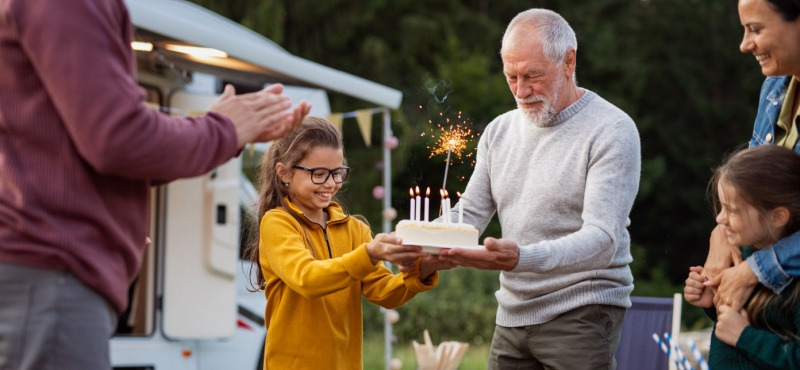 Girl presenting birthday cake with candles to elderly man outside.